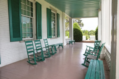 An inviting porch featuring several green rocking chairs and benches lined along the side of a building with white brick walls. Tall, green shutters complement the chairs, providing a cohesive color scheme. The porch overlooks a garden area with manicured bushes and a white picket fence, set against a serene landscape.