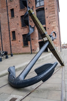 A large maritime anchor is positioned against a historical brick building with a distinct industrial design featuring large windows and a spiral staircase. The anchor is resting on a paved area with some bicycles visible in the background.