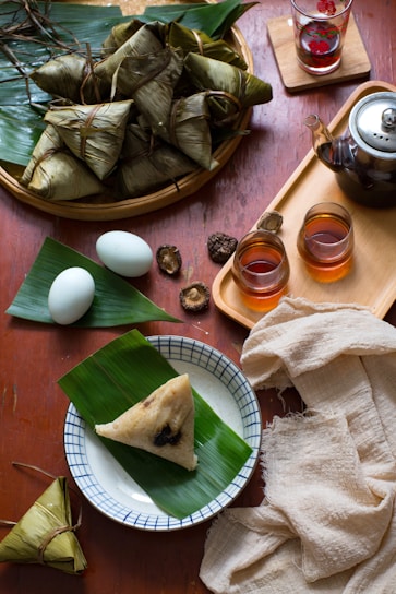 An assortment of food items, including wrapped parcels of banana leaves and a single parcel on a plate, is arranged on a wooden table. Nearby, there are two white eggs, some whole dried ingredients, and a teapot with glasses containing a warm beverage. The table is neatly organized with natural fabrics and a tray.