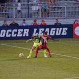 Two soccer players, one in a neon green uniform and the other in a red and black uniform, are competing for the ball on a grassy field. Behind them, a crowd of spectators is seated in a stadium with metal railings. The word 'Soccer League' is partially visible on a large banner behind the players.