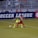 Two soccer players, one in a neon green uniform and the other in a red and black uniform, are competing for the ball on a grassy field. Behind them, a crowd of spectators is seated in a stadium with metal railings. The word 'Soccer League' is partially visible on a large banner behind the players.