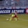 Two soccer players, one in a neon green uniform and the other in a red and black uniform, are competing for the ball on a grassy field. Behind them, a crowd of spectators is seated in a stadium with metal railings. The word 'Soccer League' is partially visible on a large banner behind the players.