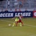 Two soccer players, one in a neon green uniform and the other in a red and black uniform, are competing for the ball on a grassy field. Behind them, a crowd of spectators is seated in a stadium with metal railings. The word 'Soccer League' is partially visible on a large banner behind the players.