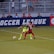 Two soccer players, one in a neon green uniform and the other in a red and black uniform, are competing for the ball on a grassy field. Behind them, a crowd of spectators is seated in a stadium with metal railings. The word 'Soccer League' is partially visible on a large banner behind the players.
