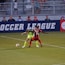 Two soccer players, one in a neon green uniform and the other in a red and black uniform, are competing for the ball on a grassy field. Behind them, a crowd of spectators is seated in a stadium with metal railings. The word 'Soccer League' is partially visible on a large banner behind the players.