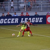 Two soccer players, one in a neon green uniform and the other in a red and black uniform, are competing for the ball on a grassy field. Behind them, a crowd of spectators is seated in a stadium with metal railings. The word 'Soccer League' is partially visible on a large banner behind the players.