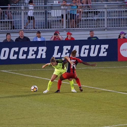 Two soccer players, one in a neon green uniform and the other in a red and black uniform, are competing for the ball on a grassy field. Behind them, a crowd of spectators is seated in a stadium with metal railings. The word 'Soccer League' is partially visible on a large banner behind the players.