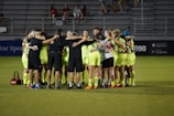 A group of soccer players in neon yellow uniforms are huddled together on a field. They are forming a tight circle, with their arms around each other, suggesting teamwork and unity. The setting appears to be a stadium during an evening match, with empty bleachers visible in the background.