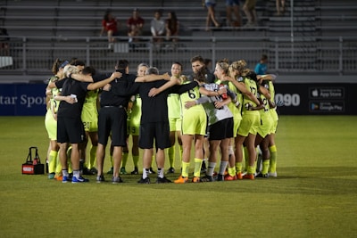 A group of soccer players in neon yellow uniforms are huddled together on a field. They are forming a tight circle, with their arms around each other, suggesting teamwork and unity. The setting appears to be a stadium during an evening match, with empty bleachers visible in the background.