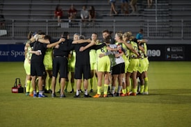 A group of soccer players in neon yellow uniforms are huddled together on a field. They are forming a tight circle, with their arms around each other, suggesting teamwork and unity. The setting appears to be a stadium during an evening match, with empty bleachers visible in the background.