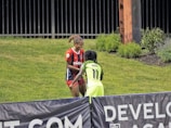 Two individuals are interacting on a grassy field. One is wearing a red and blue sports outfit with 'ProChain' visible, and the other is in a bright yellow uniform with the name 'Addo' and the number 11. They appear to be playing a sport, possibly soccer, as they are near a marked boundary. A fence and some vegetation are visible in the background.
