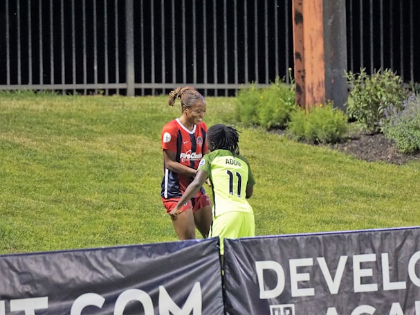 Two individuals are interacting on a grassy field. One is wearing a red and blue sports outfit with 'ProChain' visible, and the other is in a bright yellow uniform with the name 'Addo' and the number 11. They appear to be playing a sport, possibly soccer, as they are near a marked boundary. A fence and some vegetation are visible in the background.