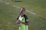 A person wearing a bright green soccer jersey is standing on a grass field. They are holding a red and black water bottle in their left hand. The jersey has a Microsoft logo and a patch on the sleeve. The background shows a grassy area with a white boundary line.