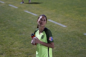 A person wearing a bright green soccer jersey is standing on a grass field. They are holding a red and black water bottle in their left hand. The jersey has a Microsoft logo and a patch on the sleeve. The background shows a grassy area with a white boundary line.