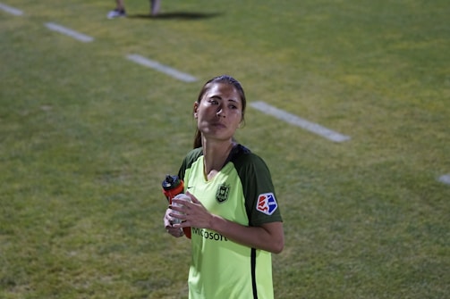 A person wearing a bright green soccer jersey is standing on a grass field. They are holding a red and black water bottle in their left hand. The jersey has a Microsoft logo and a patch on the sleeve. The background shows a grassy area with a white boundary line.