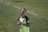 A person wearing a bright green soccer jersey is standing on a grass field. They are holding a red and black water bottle in their left hand. The jersey has a Microsoft logo and a patch on the sleeve. The background shows a grassy area with a white boundary line.