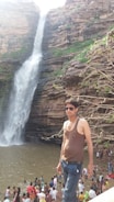 A person wearing sunglasses and a vest is standing near a waterfall with several people gathered at the base. The waterfall flows down a rocky cliff surrounded by greenery.