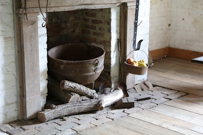 A rustic fireplace with a large metal pot sits on a bed of bricks. Several logs are stacked nearby, providing a sense of warmth and coziness. A metal hook holds a basket filled with apples and a few bananas. The setting features exposed brickwork and wooden floors, adding to the old-world charm.