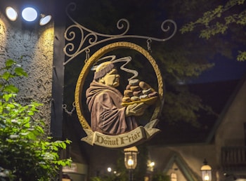 An illuminated sign hanging outside depicts a friar holding a tray piled with donuts. The sign reads 'The Donut Friar' and is artfully crafted with a circular design and ornamental ironwork. The backdrop is nighttime with ambient light from a nearby lamp post casting shadows.