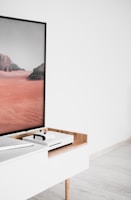 A modern interior setting featuring a white entertainment console with wooden accents. On the console, there is a gaming console with a controller on top. A large framed picture of a desert landscape with red sand and rocky formations is positioned next to the console.