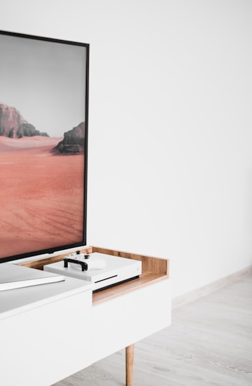 A modern interior setting featuring a white entertainment console with wooden accents. On the console, there is a gaming console with a controller on top. A large framed picture of a desert landscape with red sand and rocky formations is positioned next to the console.