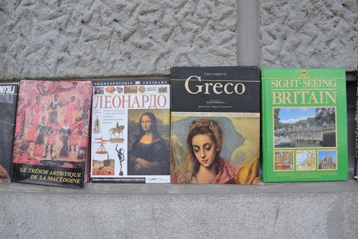 A row of books with varying covers sits against a textured stone wall. Each book has an identifiable theme or subject, such as art history or travel. The covers display artwork and text in different languages including French, Russian, and English.