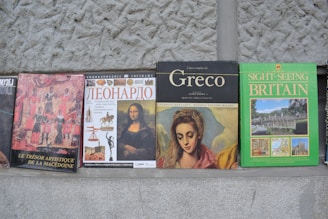 A row of books with varying covers sits against a textured stone wall. Each book has an identifiable theme or subject, such as art history or travel. The covers display artwork and text in different languages including French, Russian, and English.