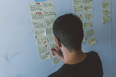 A person is organizing or affixing small sticky notes on a whiteboard. The notes contain various handwritten texts and some have small red dots on them. The person has short dark hair and is focusing on the task.