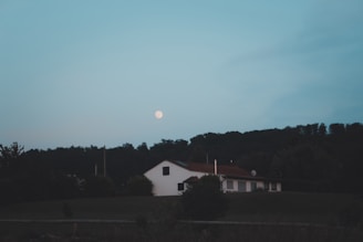 A serene evening landscape with a white house situated on a hill, surrounded by trees. The moon is visible in the sky, casting a gentle glow over the scene.