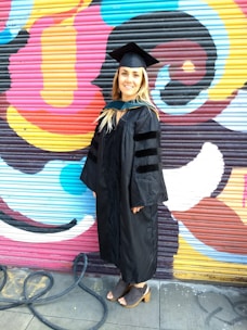 A person wearing a graduation gown and cap stands in front of a colorful, graffiti-covered wall. The background features vibrant patterns and swirling shapes in shades of pink, yellow, blue, and black. The person is smiling and appears to be celebrating a significant achievement.