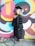 A person wearing a graduation gown and cap stands in front of a colorful, graffiti-covered wall. The background features vibrant patterns and swirling shapes in shades of pink, yellow, blue, and black. The person is smiling and appears to be celebrating a significant achievement.