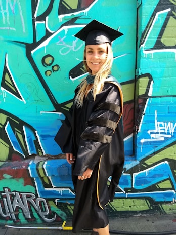 A person wearing a black graduation gown and cap stands in front of a colorful graffiti-covered wall. The wall features abstract shapes and patterns in shades of blue, green, and black with hints of white and red. The person is smiling, indicating a celebratory mood.