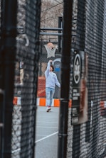 A young person wearing a white hoodie and jeans is playing basketball on an outdoor court. The scene is framed through a chain-link fence, capturing the motion of the player in action with a basketball just entering the hoop. The court features an orange and white barrier, and bare tree branches are visible in the background, suggesting a colder season.