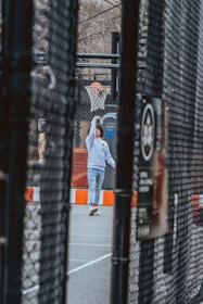 A young person wearing a white hoodie and jeans is playing basketball on an outdoor court. The scene is framed through a chain-link fence, capturing the motion of the player in action with a basketball just entering the hoop. The court features an orange and white barrier, and bare tree branches are visible in the background, suggesting a colder season.