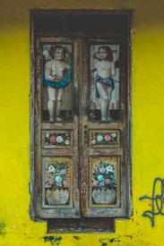 A wooden door featuring paintings of two cherubic figures and floral arrangements set against a vibrant yellow wall. The door has a weathered appearance with faded colors on the panels, which depict a child-like angelic figure on each side, surrounded by detailed and colorful floral designs. The wall surrounding the door has graffiti scribbled on the right side.