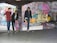 Three young men with skateboards are gathered in an urban environment filled with colorful graffiti. They stand on a concrete floor, and one of them is holding a bright pink skateboard. The walls and columns are covered in vibrant street art, creating a lively and edgy backdrop.