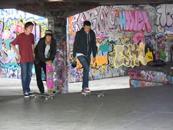 Three young men with skateboards are gathered in an urban environment filled with colorful graffiti. They stand on a concrete floor, and one of them is holding a bright pink skateboard. The walls and columns are covered in vibrant street art, creating a lively and edgy backdrop.