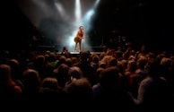 A performer stands on stage holding a guitar, illuminated by a spotlight, surrounded by smoky effects. There is a large crowd watching, with people raising their hands and taking photos.