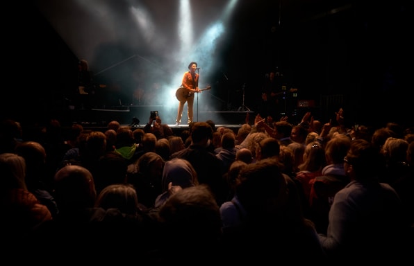 A performer stands on stage holding a guitar, illuminated by a spotlight, surrounded by smoky effects. There is a large crowd watching, with people raising their hands and taking photos.