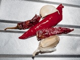 Several dried red chili peppers and two cloves of garlic are arranged on a metallic, textured surface. The chilies have a shiny appearance, suggesting they are dry and slightly wrinkled. The garlic cloves are peeled, revealing their smooth, off-white surface.
