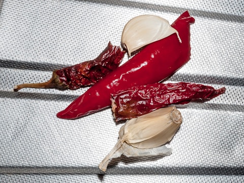 Several dried red chili peppers and two cloves of garlic are arranged on a metallic, textured surface. The chilies have a shiny appearance, suggesting they are dry and slightly wrinkled. The garlic cloves are peeled, revealing their smooth, off-white surface.