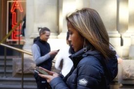 A woman in a black puffer jacket is focused intently on her smartphone. In the background, a young man is also looking at his phone while standing on stone steps. The scene includes a bright, red neon 'SALE' sign and architectural elements of a building.