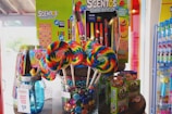 A colorful display featuring various creative sets and candy. Large rainbow lollipops are placed in a jar filled with multicolored candy. A variety of scented markers and pencils, labeled 'Scentos,' are on display in vibrant packaging. A blue mason jar with a sign reading 'Ducky Pens' is also visible.