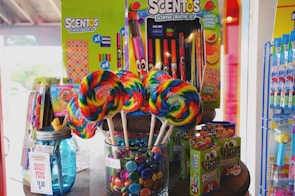 A colorful display featuring various creative sets and candy. Large rainbow lollipops are placed in a jar filled with multicolored candy. A variety of scented markers and pencils, labeled 'Scentos,' are on display in vibrant packaging. A blue mason jar with a sign reading 'Ducky Pens' is also visible.