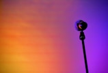 A lone microphone stands against a vibrant, gradient background transitioning from warm orange to deep purple, creating a dramatic and artistic effect.