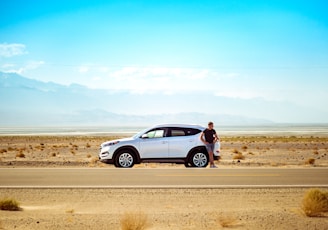 man standing beside white SUV near concrete road under blue sky at daytime