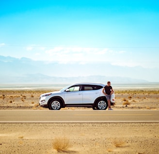 man standing beside white SUV near concrete road under blue sky at daytime