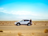 man standing beside white SUV near concrete road under blue sky at daytime
