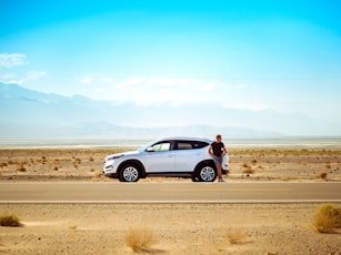 man standing beside white SUV near concrete road under blue sky at daytime