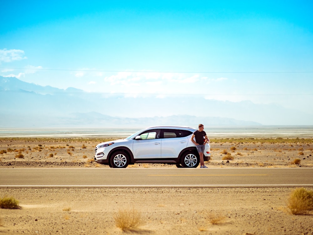 hombre de pie junto a un SUV blanco cerca de una carretera de hormigón bajo el cielo azul durante el día